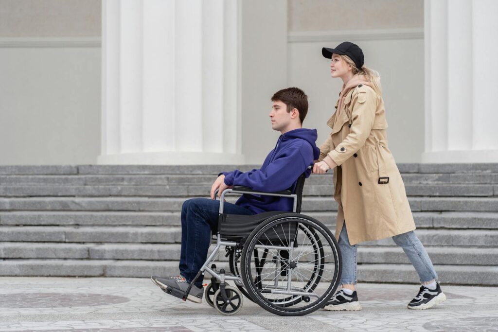 A young woman helps a man in a wheelchair around an outdoor location with white columns.