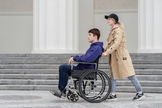 A young woman helps a man in a wheelchair around an outdoor location with white columns.